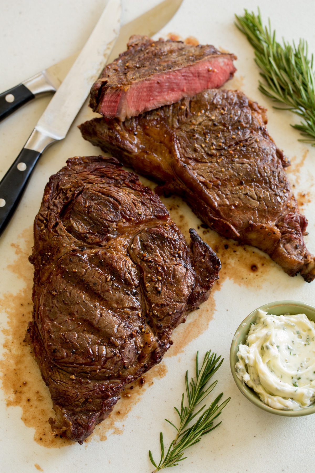 Two grilled steaks on a cutting board with knives and rosemary to the side.