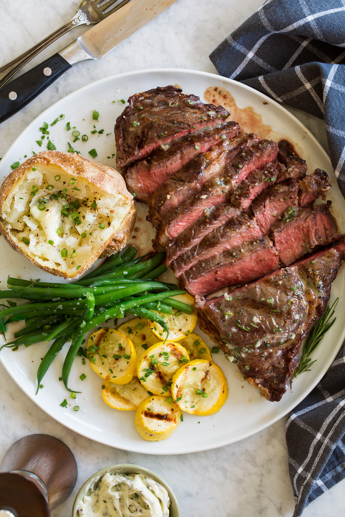 Grilled steak shown sliced on a serving plate with a side or grilled squash, green beans and a baked potato.