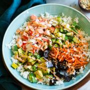 California roll sushi bowl shown in a blue bowl from a side angle. Bowl is resting on a wooden plate over a marble surface.