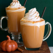 Pumpkin spice latte. Shows two servings in glass mugs one a wooden plate set over a white marble surface with a green background.