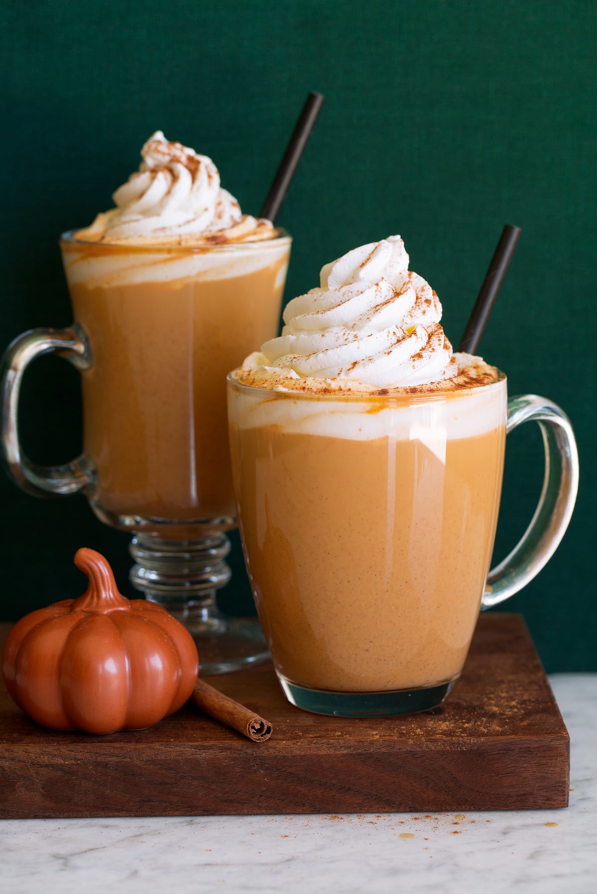 Pumpkin spice latte. Shows two servings in glass mugs one a wooden plate set over a white marble surface with a green background.