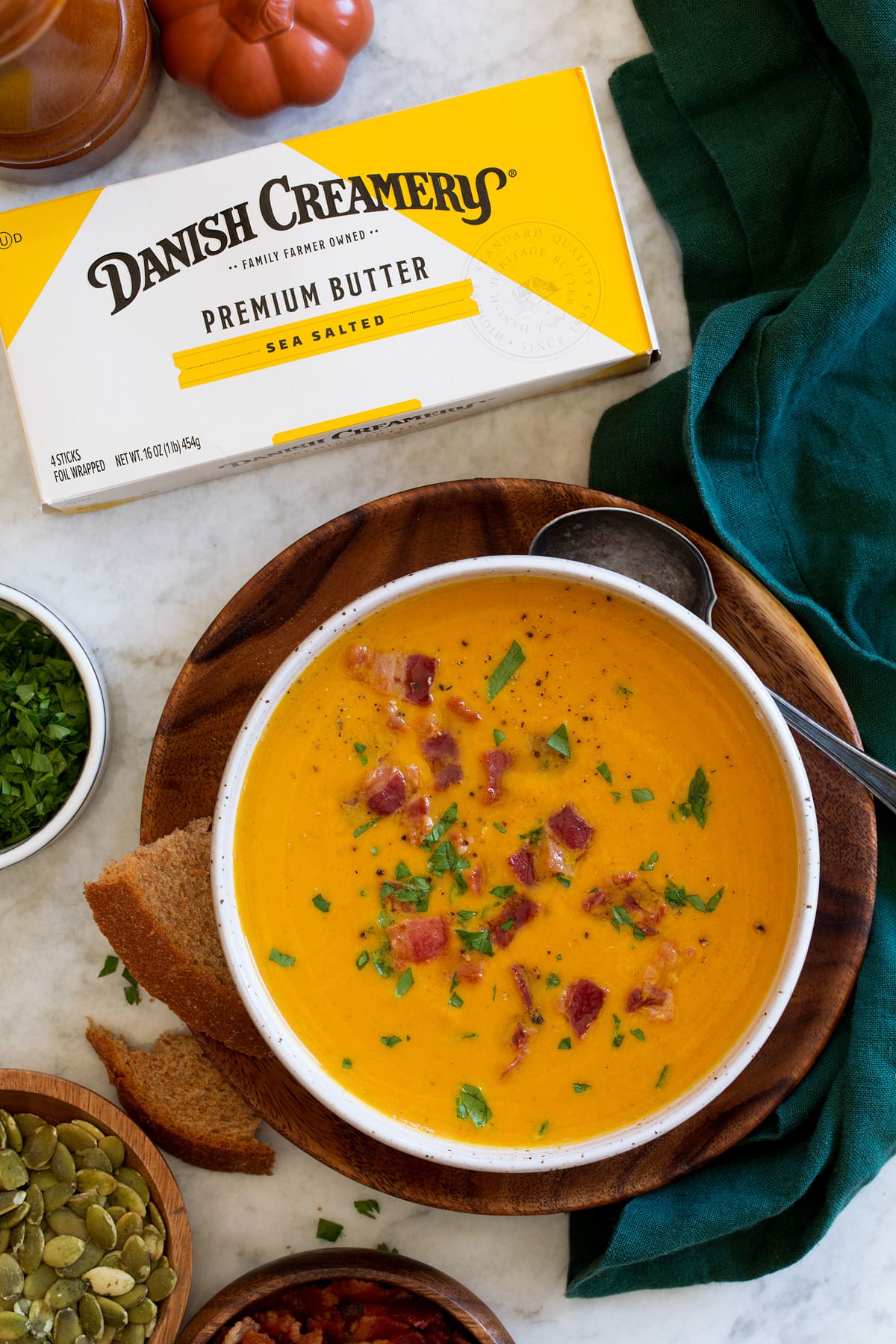 Pumpkin soup in a white bowl over a wooden plate with wheat bread and butter to the side.