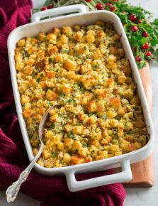 Cornbread stuffing shown in a white rectangular baking dish over a wooden platter with herbs, cranberries and a red cloth to the side.