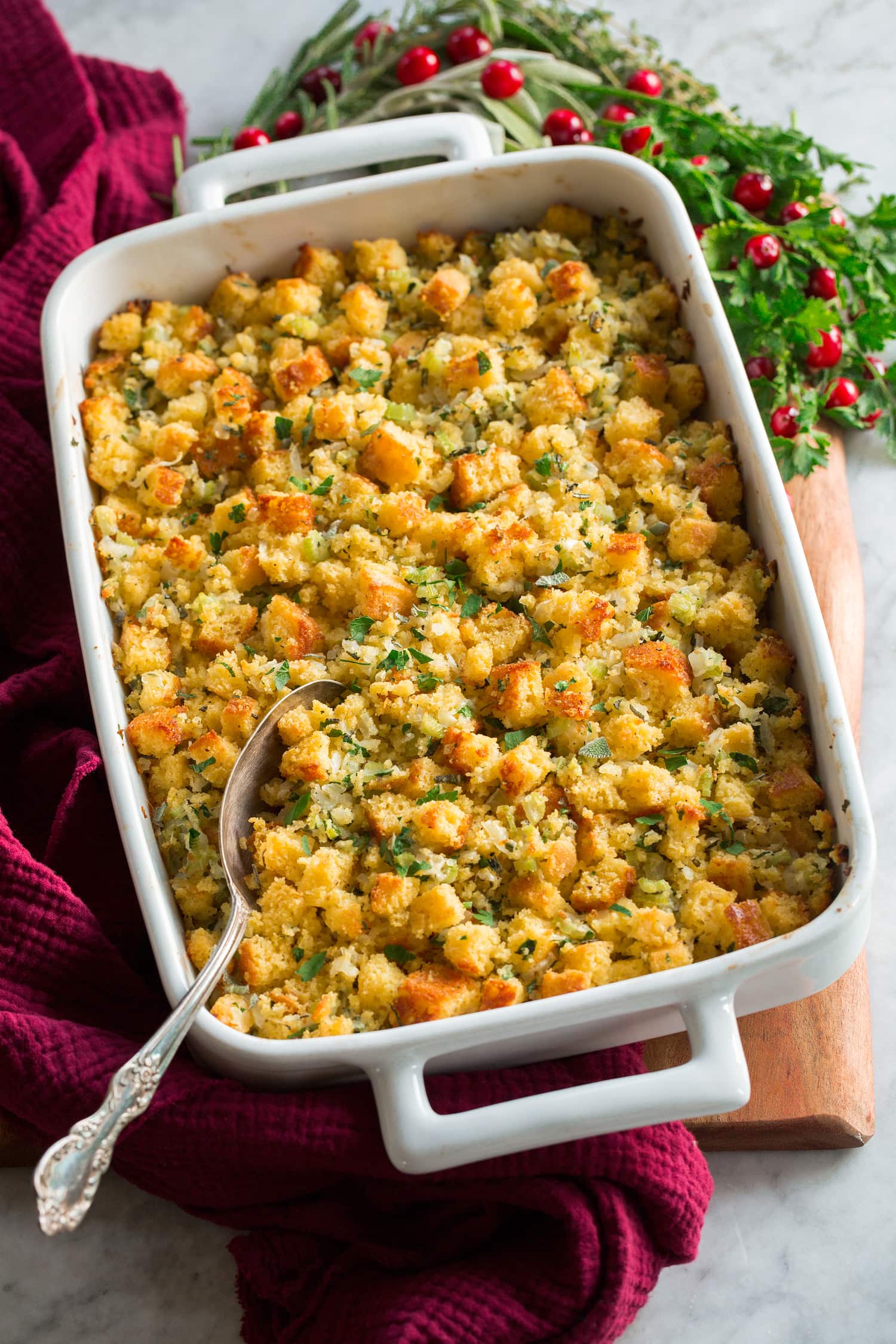 Cornbread stuffing shown in a white rectangular baking dish over a wooden platter with herbs, cranberries and a red cloth to the side.