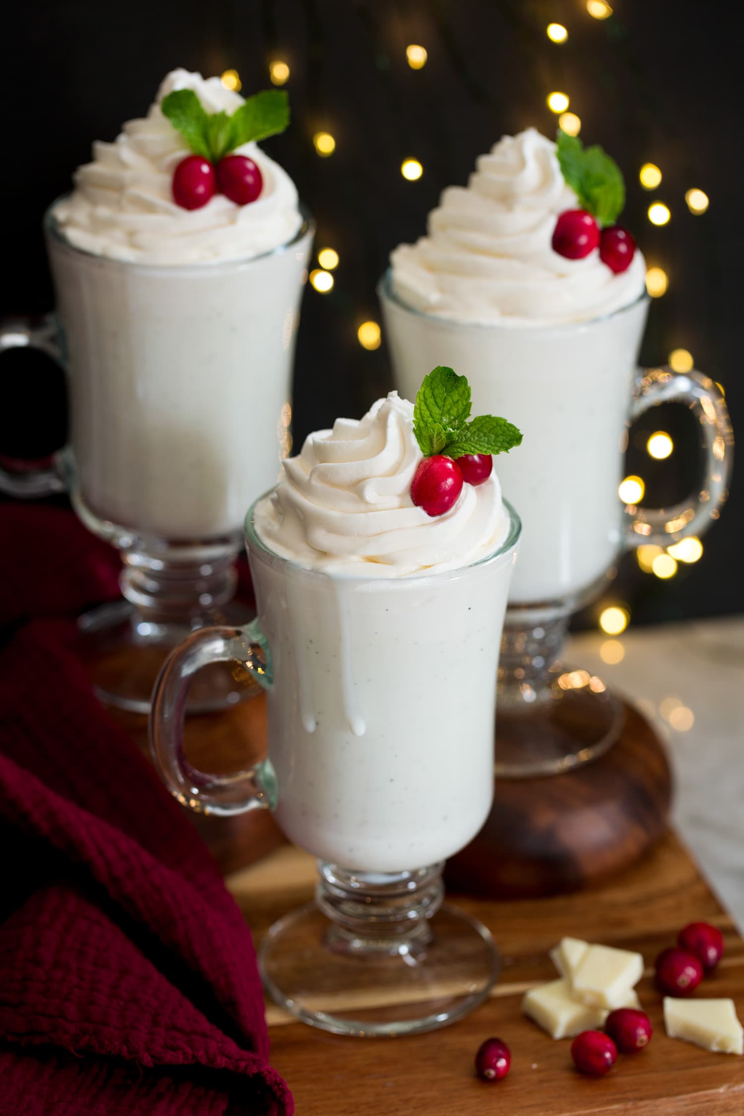 Three mugs of white hot chocolate on a wooden tray with Christmas lights in the background.