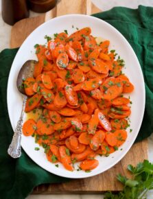 Glazed carrots shown served on a white oval platter set over a wooden tray and a green cloth.