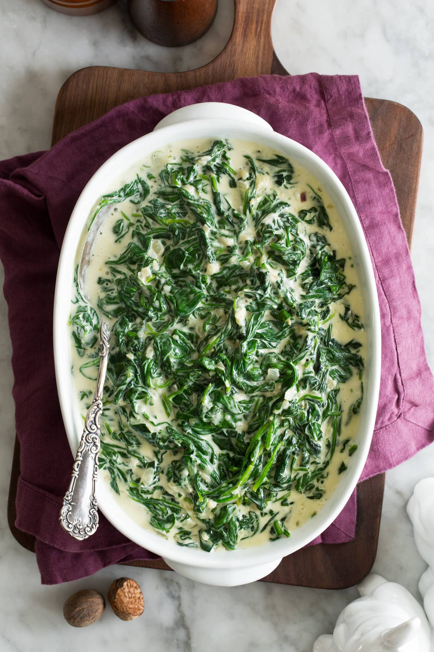 Fresh creamed spinach in a white oval serving dish set over a purple cloth and a wooden tray.