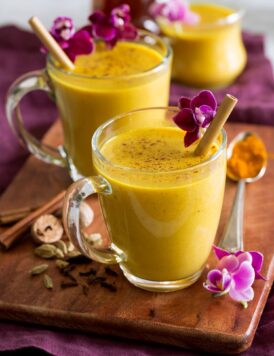 Golden milk shown in glass mugs on a wooden tray with spices and flowers to the side.