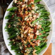 Green Bean Almondine. Sauteed green beans are shown lined up in a row on a platter with toasted almonds, crumbled fried bacon and grated parmesan are sprinkled over the top.