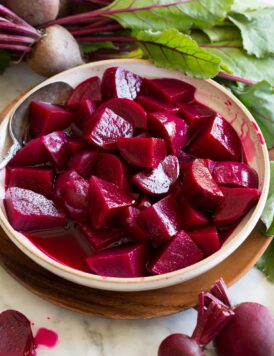 Chopped homemade pickled beets shown in a bowl surrounded by fresh beets.