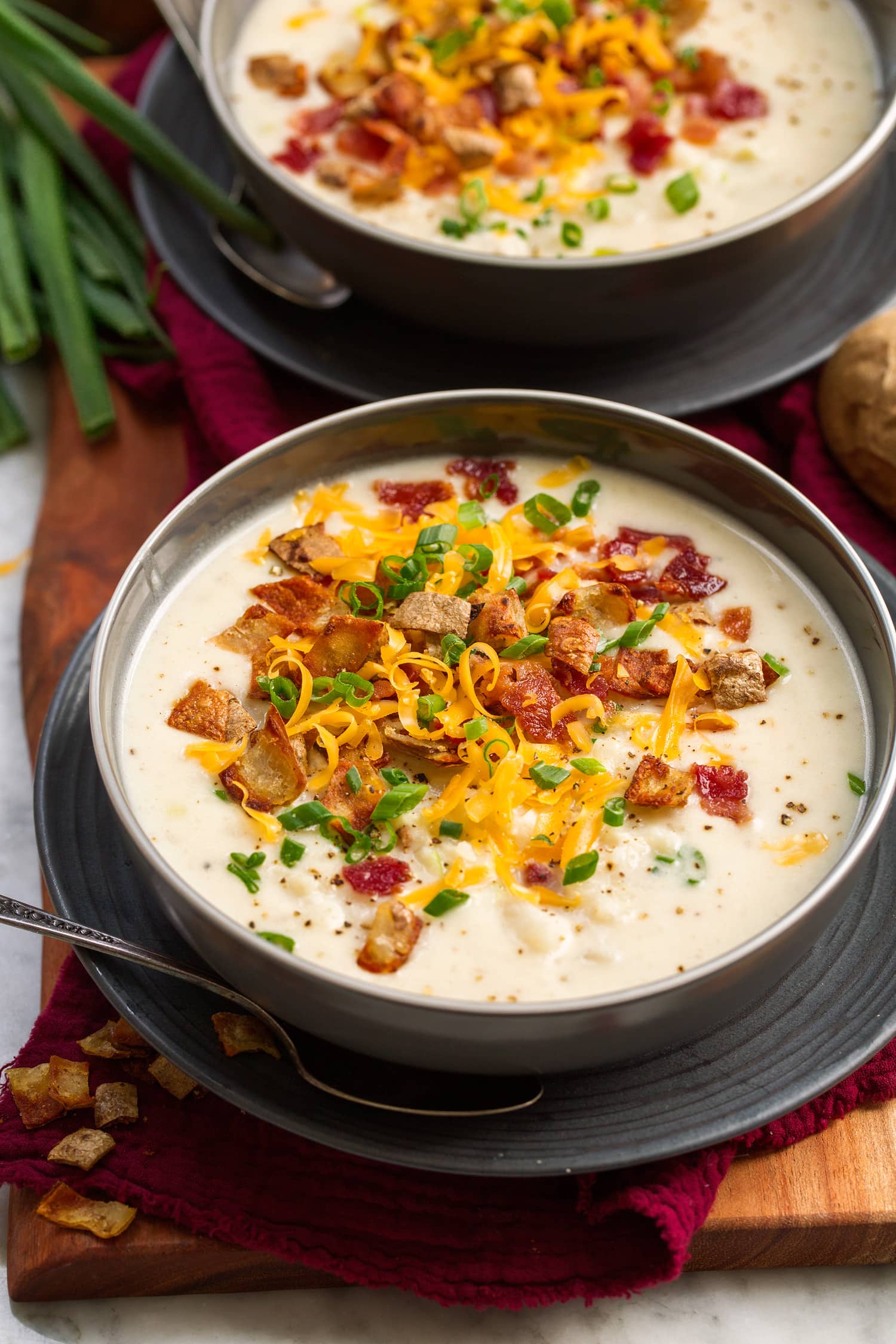 Serving of baked potato soup in a gray bowl over a gray plate. It is topped with cheddar cheese, fried potato skins, bacon and green onions.