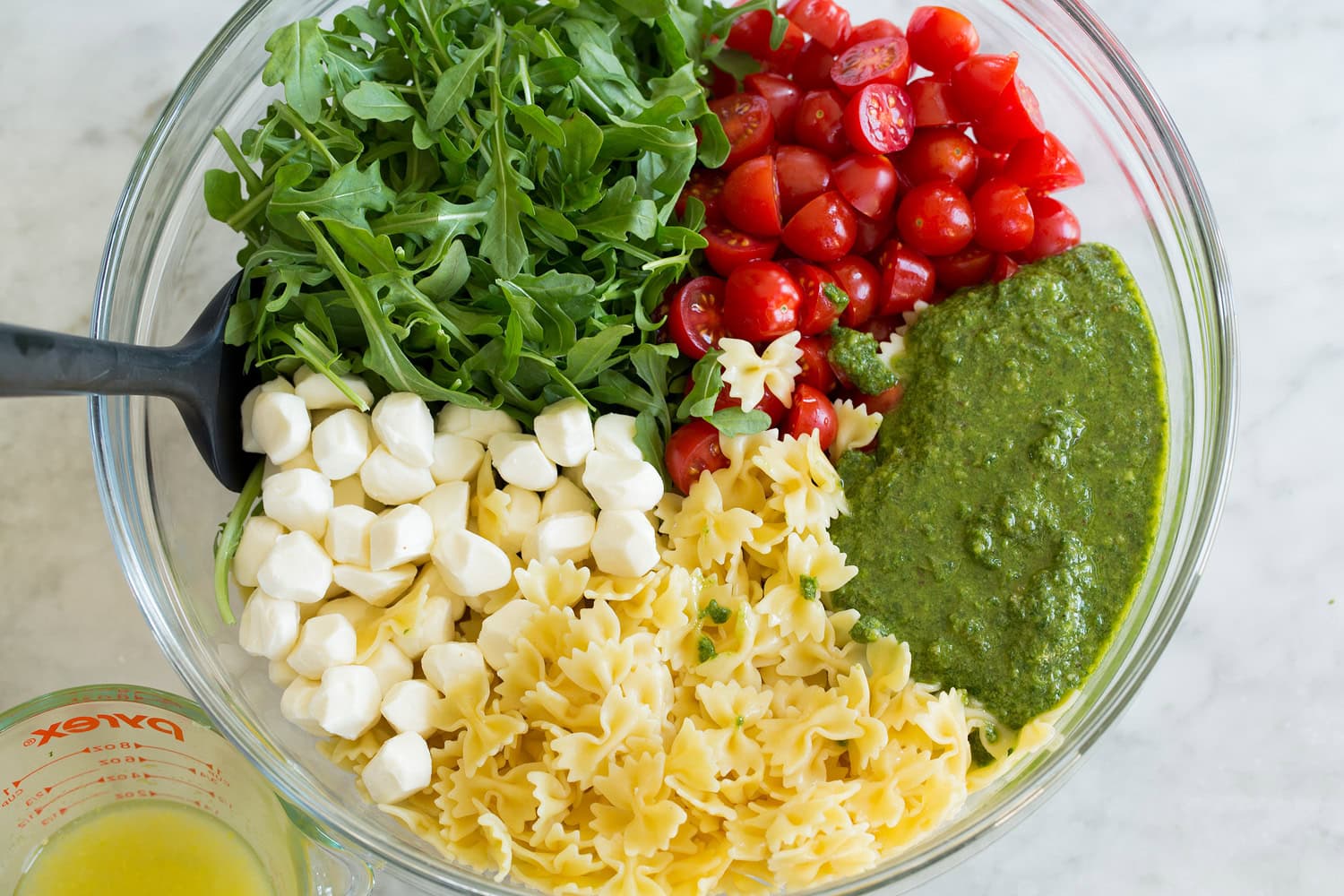 Pesto pasta salad ingredients shown before tossing salad in a glass mixing bowl.